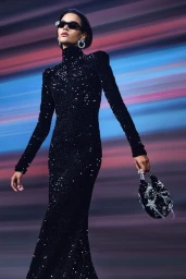 Model in black sequined turtleneck dress wearing sunglasses against a streaked colourful background holding an embellished handbag.