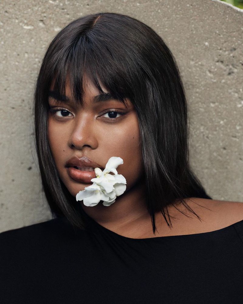Close-up portrait of Precious Lee holding a white flower in her mouth against a textured stone wall.