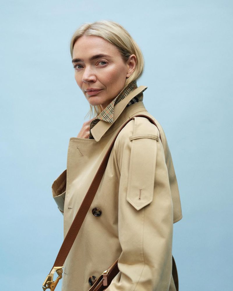 Portrait of a mature woman in a trench coat against a light blue background, looking at the camera with a neutral facial expression.