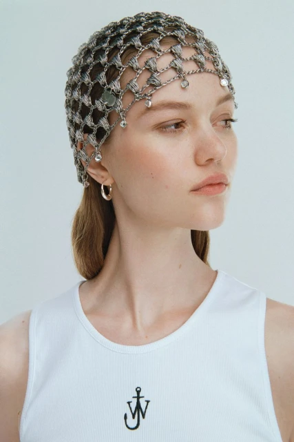 Close-up of a woman with a metallic net head covering and white tank top, neutral background, studio lighting, silver hoop earrings.