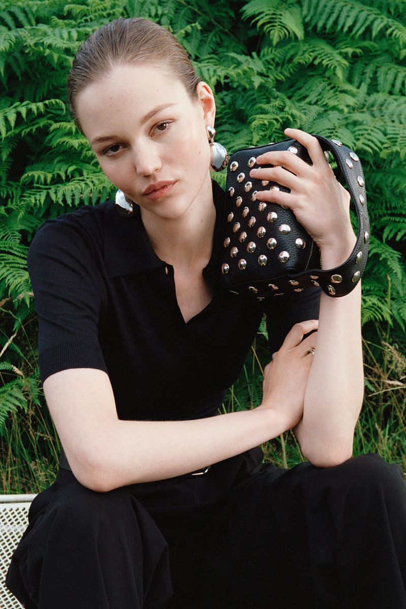 Attractive woman poses in black outfit holding black studded purse in front of lush green fern background in outdoor setting.