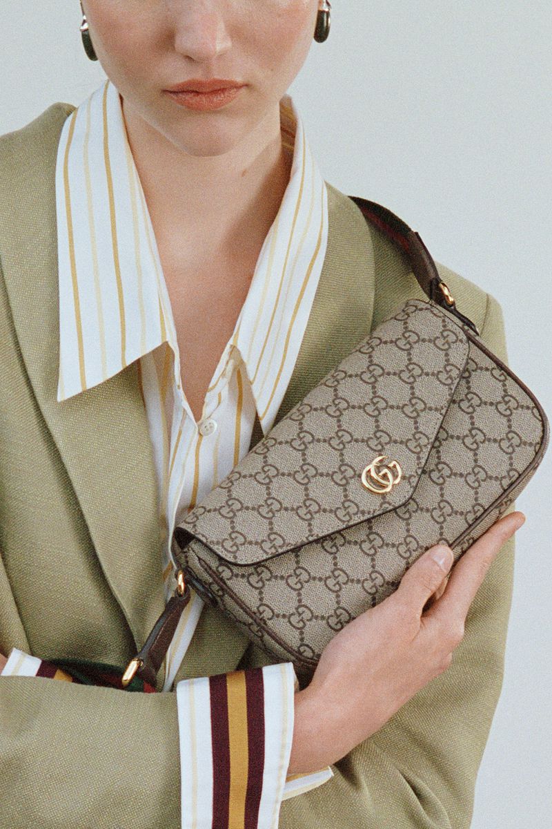 Fashion shot featuring a light-skinned woman in a striped shirt and blazer holding a Gucci bag with logo pattern.