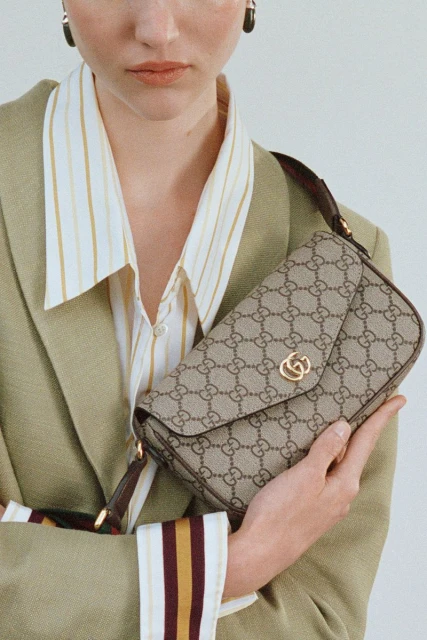 Fashion shot featuring a light-skinned woman in a striped shirt and blazer holding a Gucci bag with logo pattern.