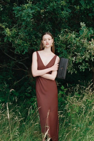Model Noor Chaltin wearing a brown slip dress in a grassy meadow holding a dark clutch bag