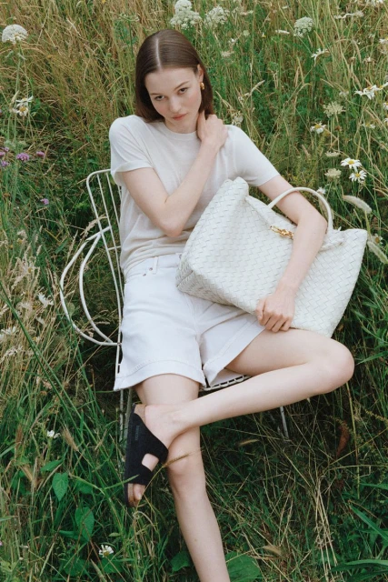 A young woman in a white t-shirt and shorts sits in a grassy field, holding a woven tote bag, posing for a fashion editorial.