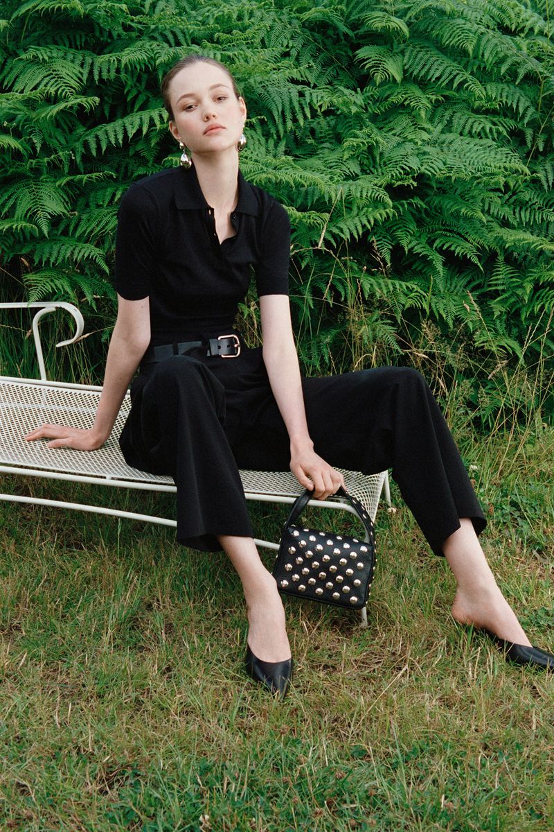 Model wearing a black polo shirt and trousers seated on a white garden bench outdoors against green ferns