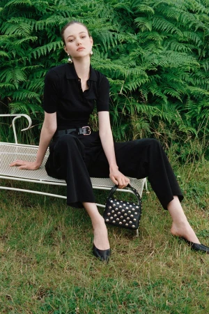 Model wearing a black polo shirt and trousers seated on a white garden bench outdoors against green ferns