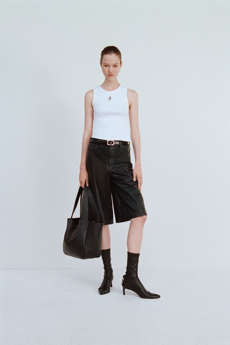 Woman posing in a white ribbed tank top, black leather culottes, black heeled sock boots, and holding a black leather handbag on a white backdrop.