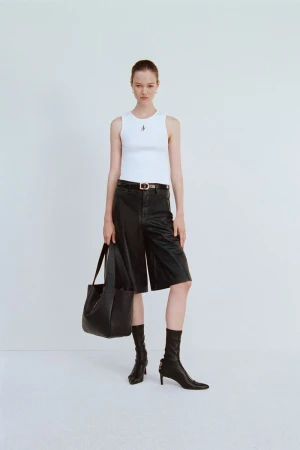 Woman posing in a white ribbed tank top, black leather culottes, black heeled sock boots, and holding a black leather handbag on a white backdrop.