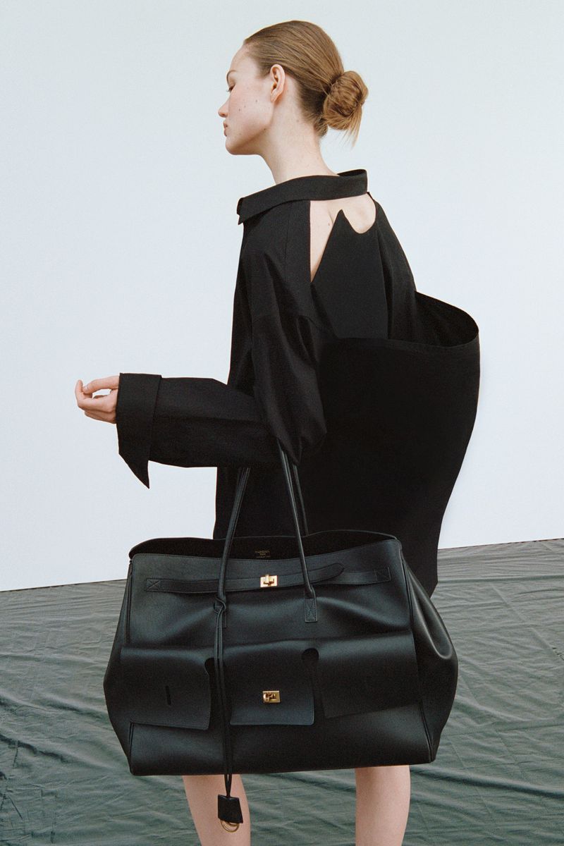 Woman in black dress carries a large black leather tote bag in a fashion editorial, studio lighting with a gray backdrop.