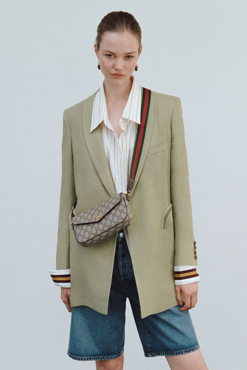 A woman poses in an olive blazer, striped shirt, and blue denim shorts with a Gucci bag and shoulder strap against a plain background.