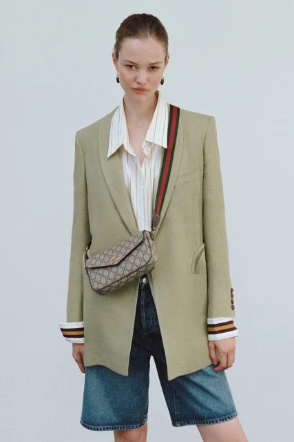 A woman poses in an olive blazer, striped shirt, and blue denim shorts with a Gucci bag and shoulder strap against a plain background.