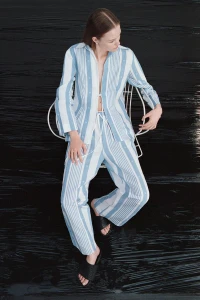 Model wearing a blue and white striped shirt and matching trousers while seated on a white wire chair against a dark glossy background.