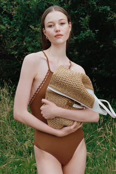 Model in brown swimsuit holding a woven straw handbag outdoors against a lush green meadow backdrop
