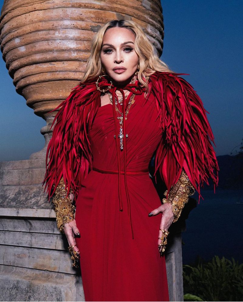Madonna in red gown with feather boa and gold jewelry, posing by stone vase against blurred background in portrait orientation.