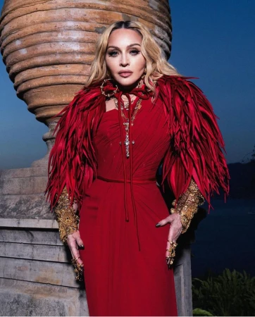 Madonna in red gown with feather boa and gold jewelry, posing by stone vase against blurred background in portrait orientation.