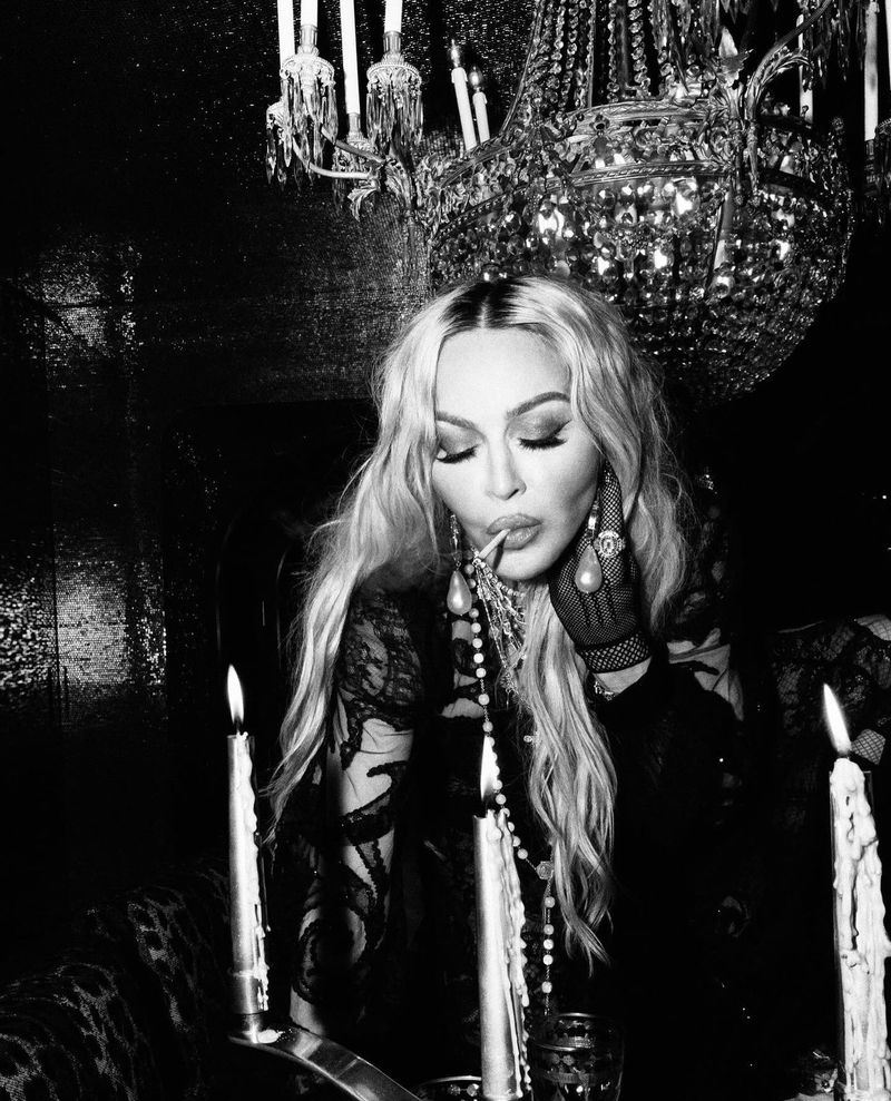 Black-and-white portrait of a blonde adult female with a cigarette, posing in front of a chandelier and lit candles in an indoor setting.
