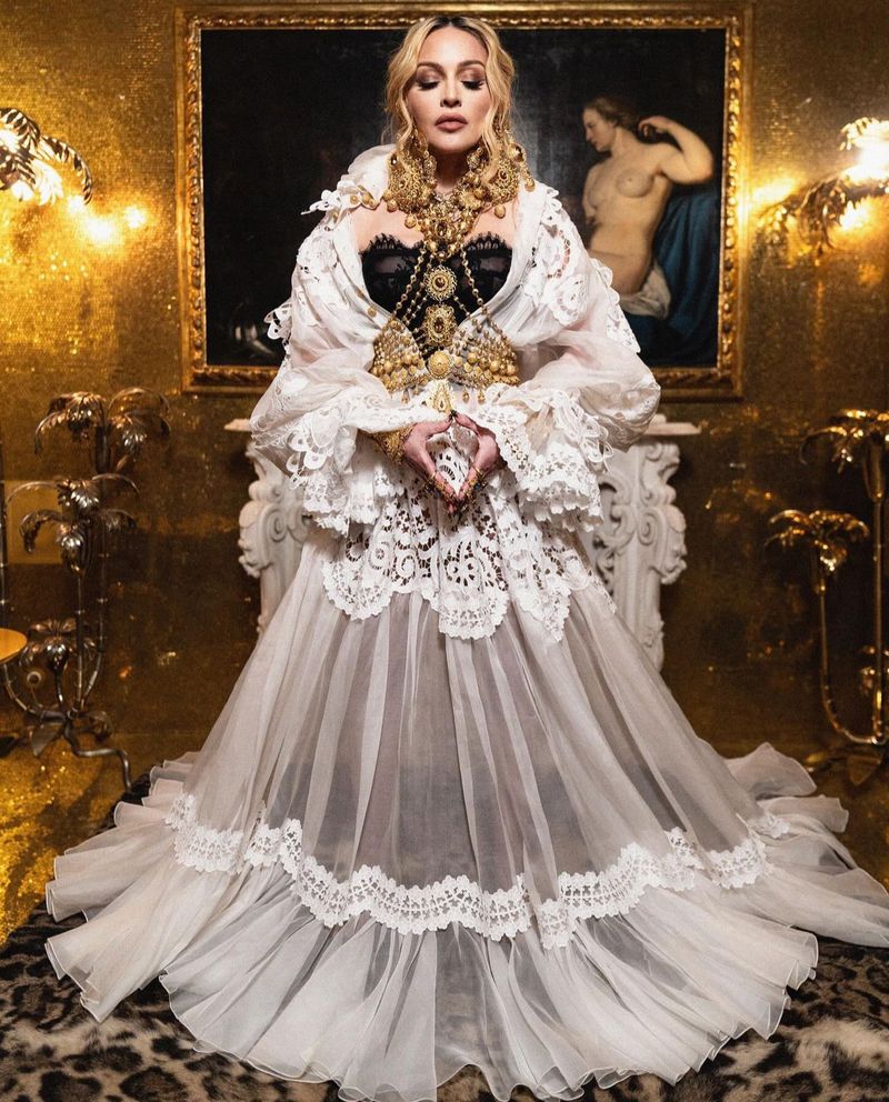 Adult female in elegant gown with lace details, gold jewelry, ornate background in an indoor environment with dramatic lighting.