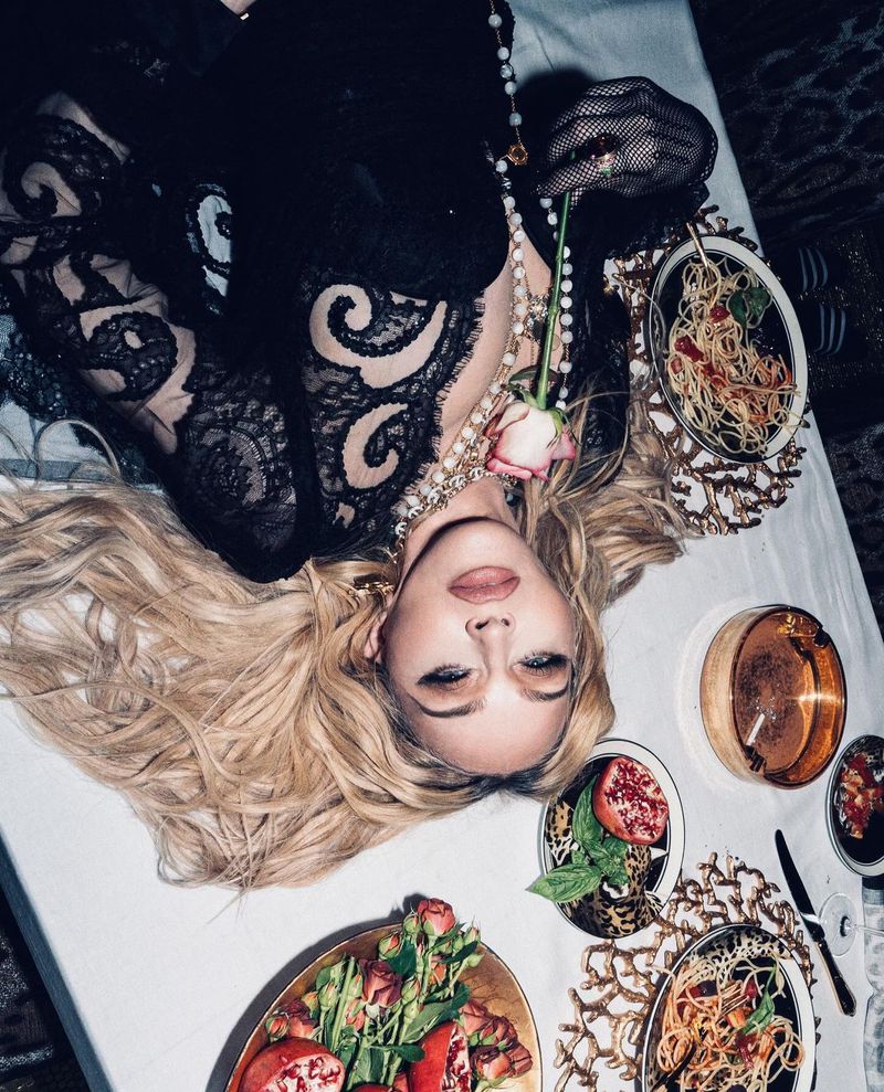 Mature adult woman with blonde hair lying on a white-tablecloth-covered table with dishes of food, wearing a lace dress.