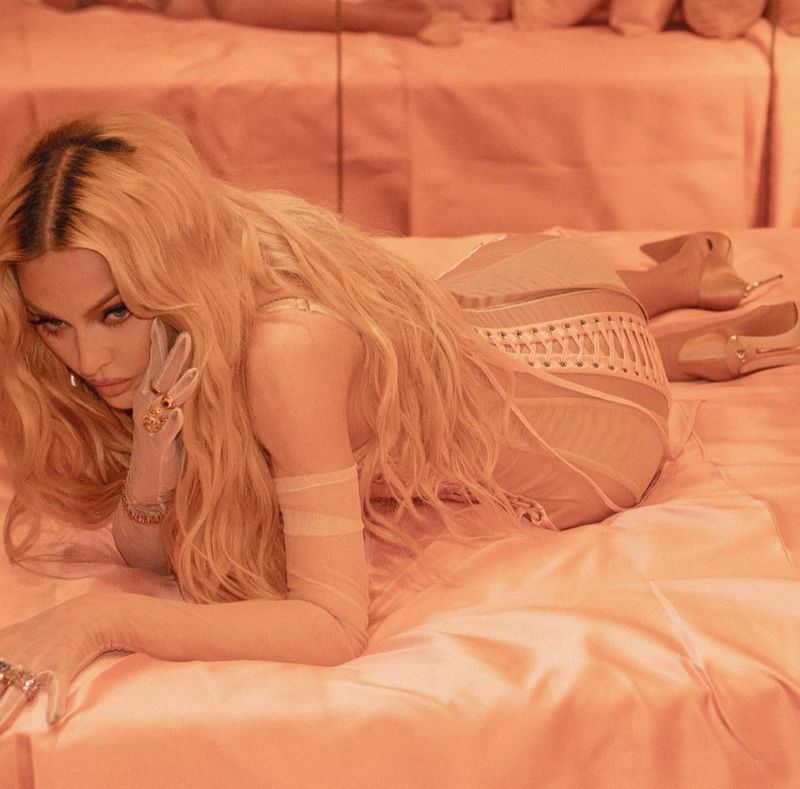 Madonna, an adult female, poses on a bed in a sheer dress and heels, with a peach-colored monochrome aesthetic in an indoor setting.