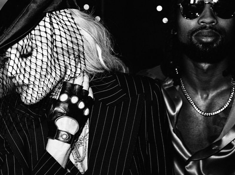 Monochrome portrait of a veiled blonde woman in a pinstripe suit and black gloves, standing next to a man in a silk shirt and chain.
