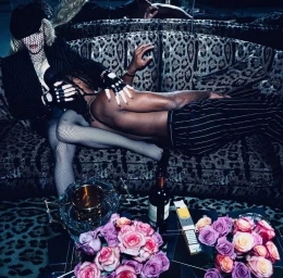 Madonna seated on a leopard print couch wearing a pinstripe jacket and fishnet veil, with another figure reclining across her lap