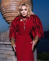 Madonna wearing a red dress with feathered shoulders and ornate gold jewelry outdoors next to a large stone urn.