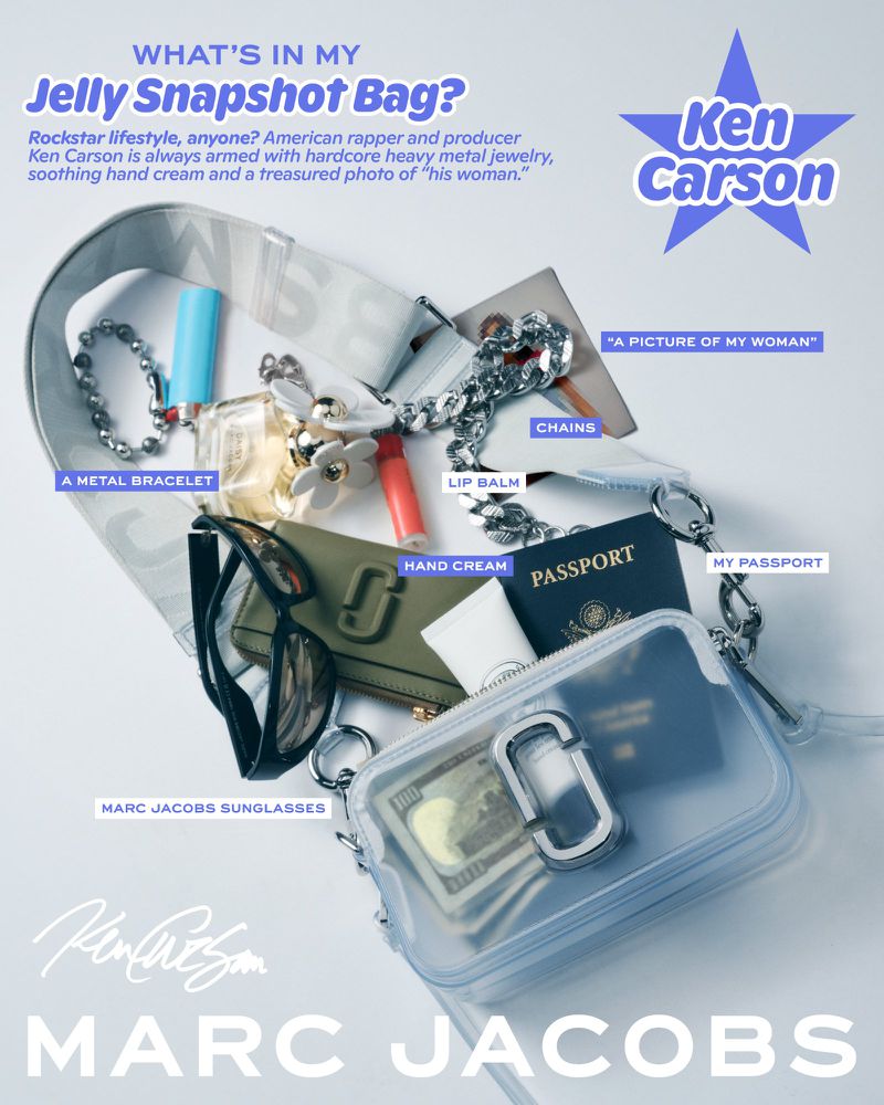 Overhead shot of a clear Marc Jacobs handbag filled with everyday items: passport, lip balm, sunglasses, hand cream, metal jewelry.