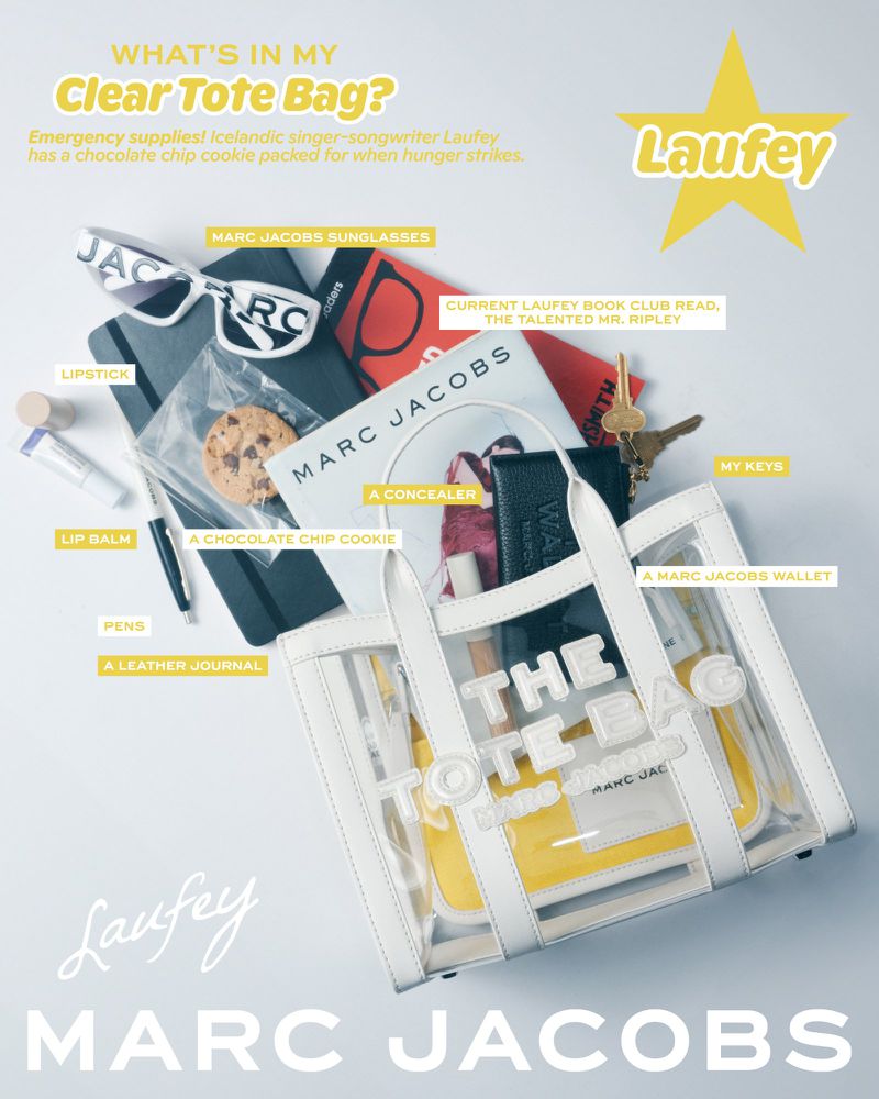 Clear tote bag by Marc Jacobs featuring sunglasses, book, wallet, keys, cosmetics, and cookie; laid flat on a white surface.