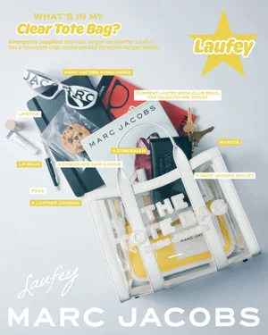 Flat lay of a clear Marc Jacobs tote bag surrounded by personal items including sunglasses, a journal, keys, and a cookie.