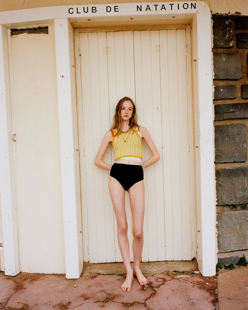 A slim, barefoot young woman with pale skin is posing in front of a closed white door with the text CLUB DE NATATION above it.