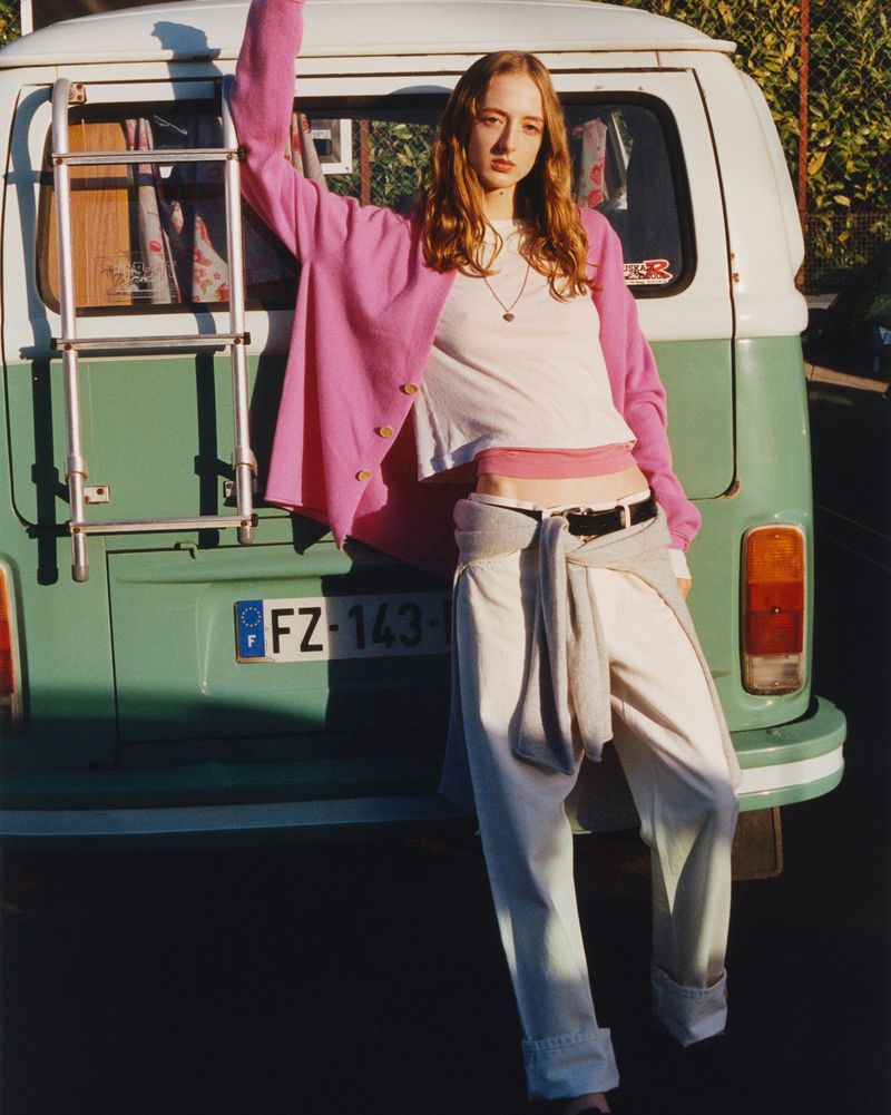 A woman leans against a green and white vintage van, wearing a pink cardigan, white top, and white pants in an outdoor setting.