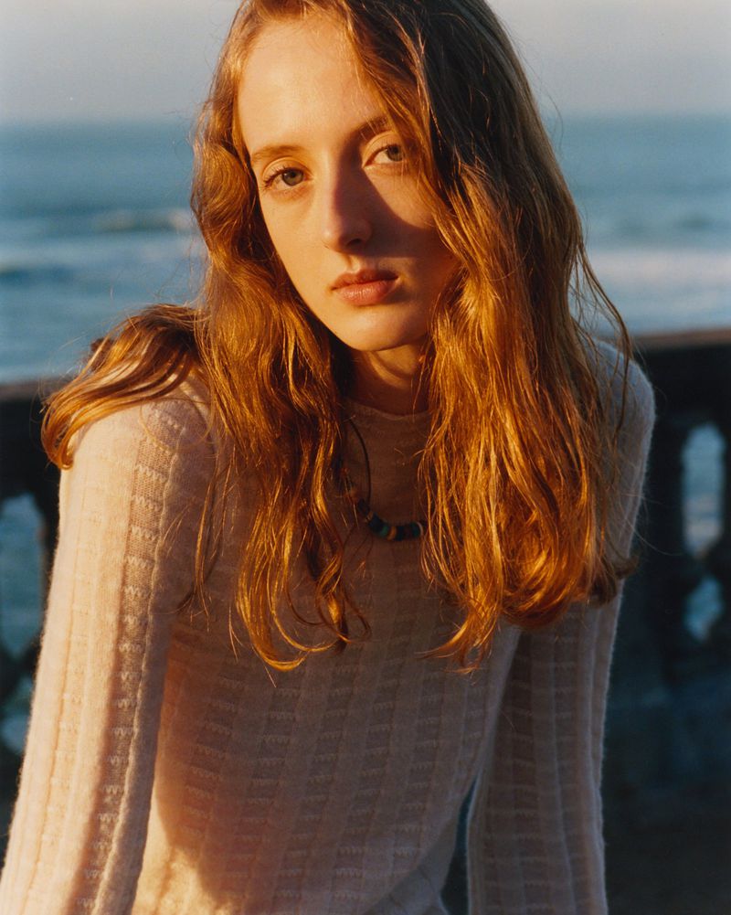 Close-up portrait of a young woman with long red hair and a light complexion, wearing a textured sweater with a sea background.