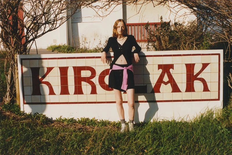 Adult female model poses in front of a large KIRIAK sign in an outdoor setting with a black jacket, black skirt, and pink belt.