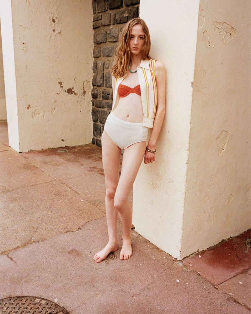 Young woman in a two-piece swimsuit and knit vest stands next to an off-white wall with stone trim in a medium shot.