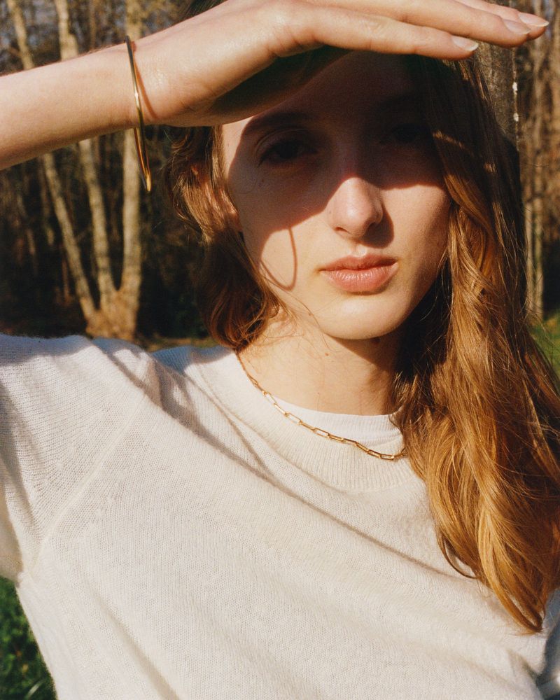A young adult woman stands outdoors in the sunlight, shielding her eyes with her hand. She wears a white top and gold chain.