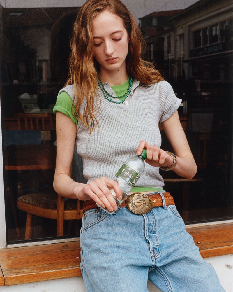 Young woman opens a water bottle while sitting on a windowsill in an outdoor setting with natural lighting and a retail storefront.