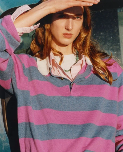 Close-up of a model in a pink and gray striped sweater shielding her eyes, creating a shadow on her face in an indoor setting.