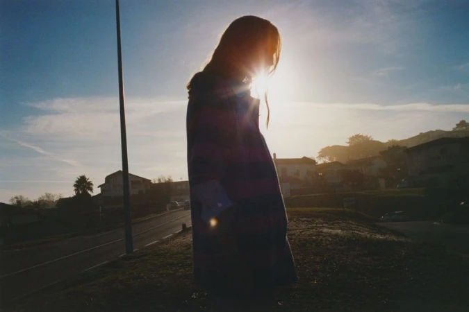 Silhouette of a person standing in a field with houses and trees in the background, illuminated by the sun, creating a bright backlight.