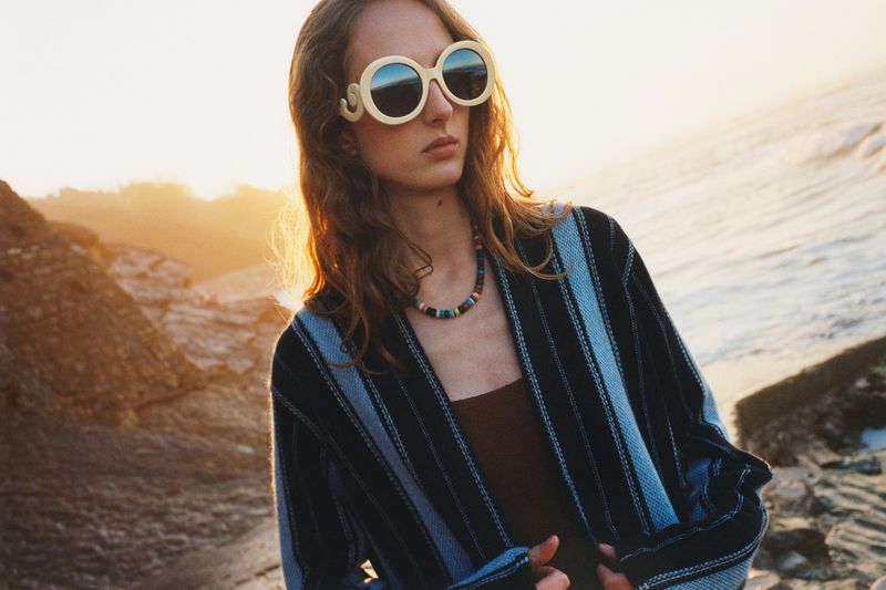 Woman wearing sunglasses and a striped jacket on a rocky beach at sunset, fashion editorial. Warm lighting and long wavy hair.