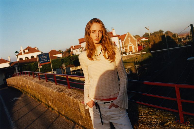 Woman in white sweater and jeans poses on bridge with town buildings in background, bathed in soft, natural light during daytime.