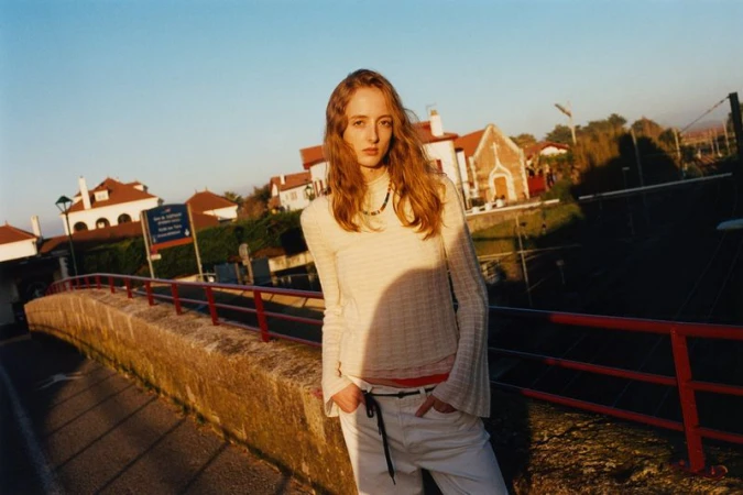 Woman in white sweater and jeans poses on bridge with town buildings in background, bathed in soft, natural light during daytime.