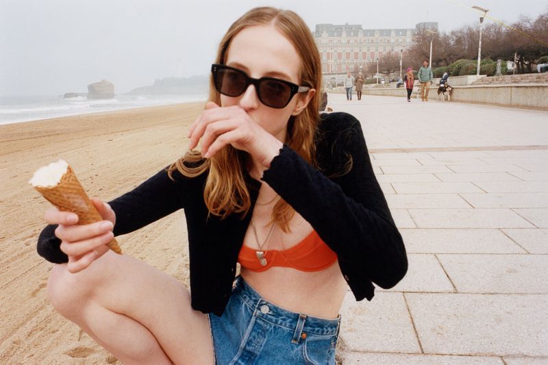 Young woman on beach enjoying ice cream cone in swimwear and sunglasses, seated on seaside promenade with ocean and city in background.