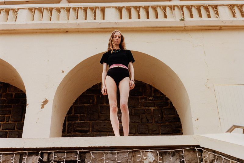 Woman stands on a wall in black crop top and high-waisted bottoms, posing against an arched stone building, bathed in soft, diffused light.