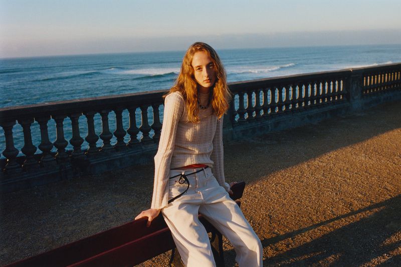 A young woman with long wavy red hair wearing casual clothes sits on a bench near a stone wall with the ocean and blue sky behind her.
