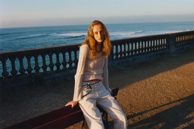 A young woman with long wavy red hair wearing casual clothes sits on a bench near a stone wall with the ocean and blue sky behind her.