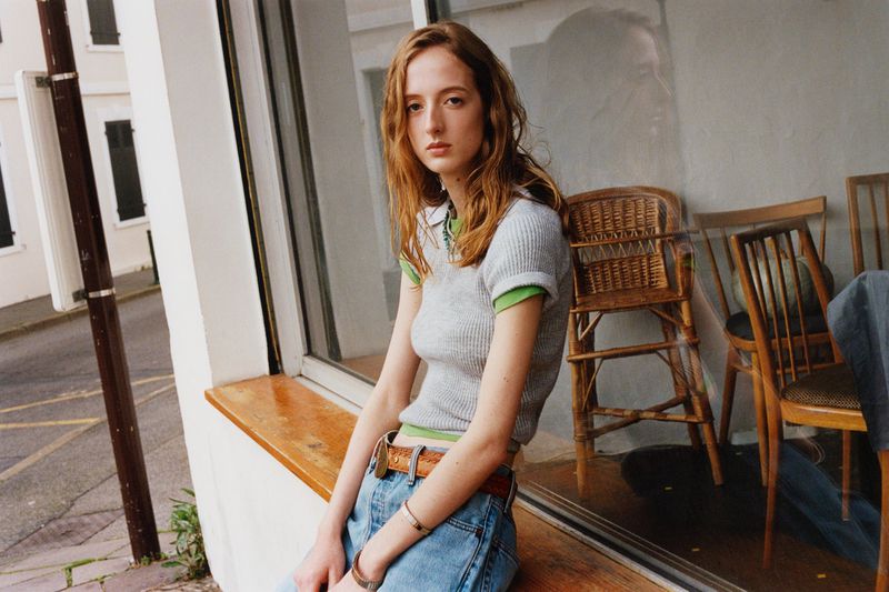 Young woman with long brown hair sitting near a window, wearing a grey top and blue jeans, with chairs and an outdoor scene visible.