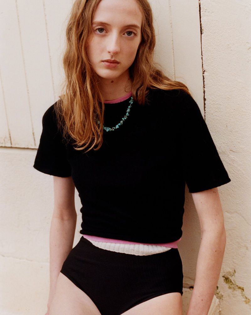Woman in black knit top and briefs poses against a cream wall, showcasing a modern, minimalist fashion aesthetic with natural light.