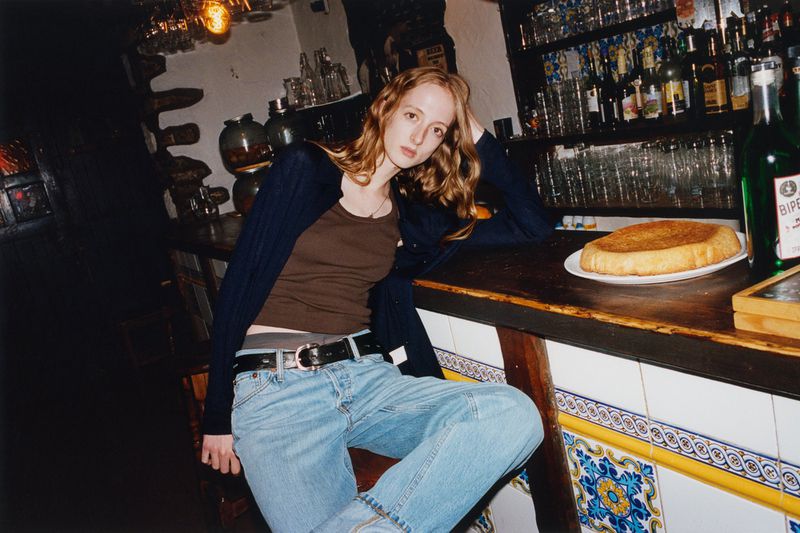 Young woman with long auburn hair sits at a bar in a brown top and blue jeans.  There's a bottle of BIPE and some food on the bar.