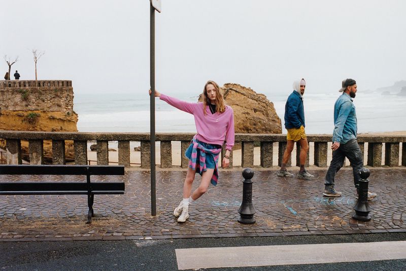 Woman poses by a pole near the ocean on a cloudy day, wearing a pink sweatshirt with two men walking behind her in the distance.